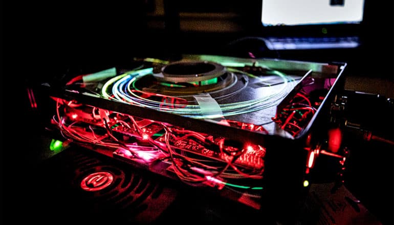 The concussion detection device sits on a table in a dark room, red lights coming from its open sides and green light coming from fibers coiled on its top