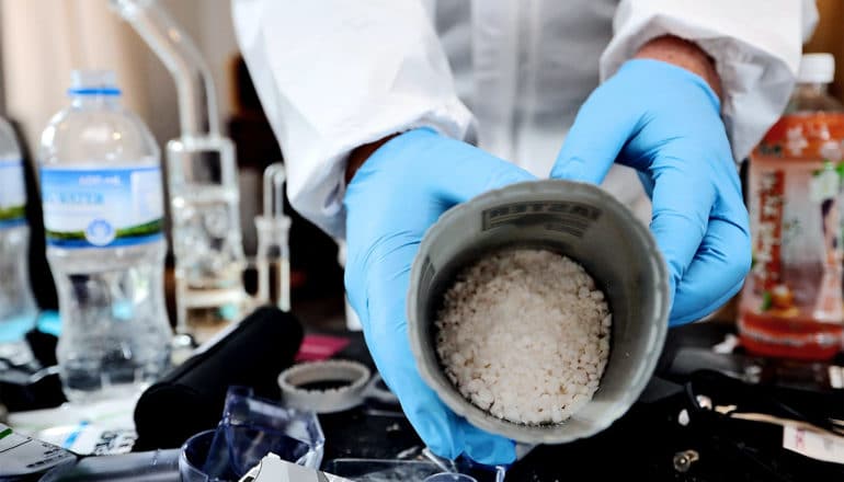 A person with white sleeves and blue gloves holds a container of small white crystals