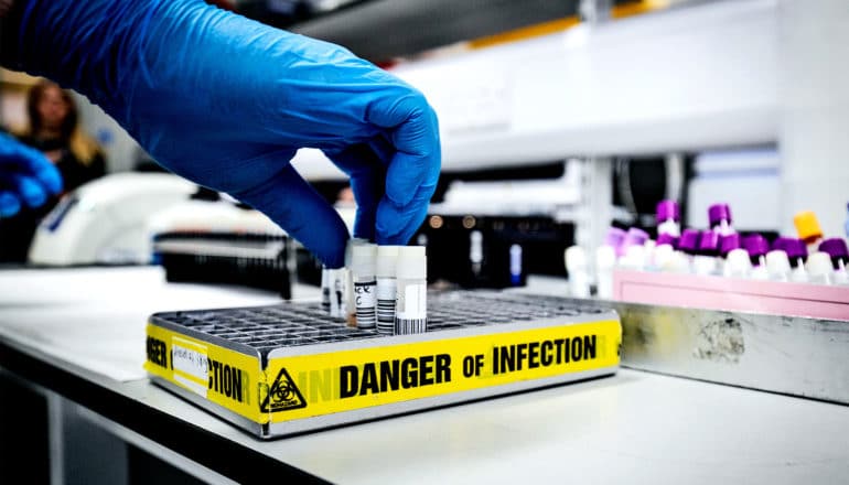 A researcher wearing blue gloves reaches for a vial of coronavirus held in a container labeled "Danger of Infection" on yellow warning tape