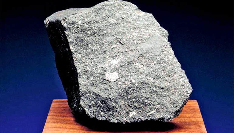 The meteorite chunk Curious Marie is light gray and sits on a wooden block against a black and blue background