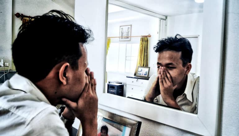A man covers his mouth and nose with his hands as he looks at himself in a mirror in a mostly white-painted home