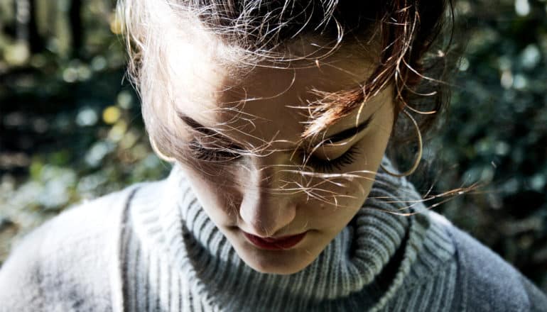 A young woman in a grey sweater walks outside while looking down, with her face in shadow and light shining on her forehead and hair