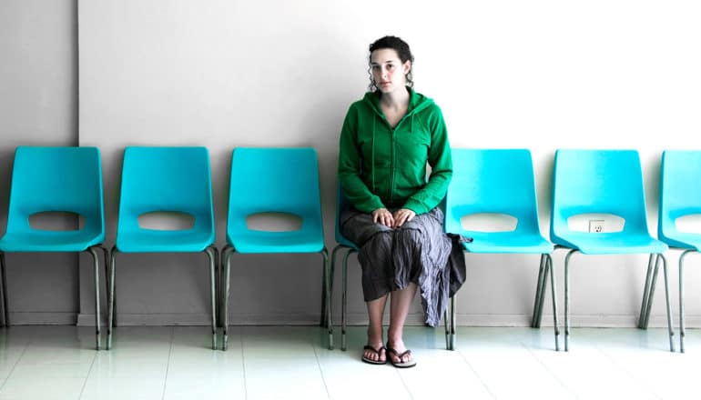 A woman in a green hoodie sits on a blue plastic chair in a doctor's waiting room