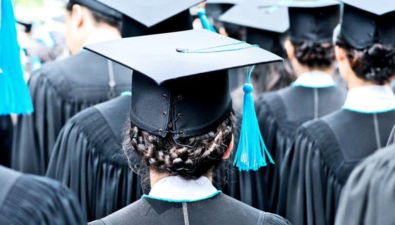 A group of college graduates in black robes and caps with blue tassels stand facing away from the camera