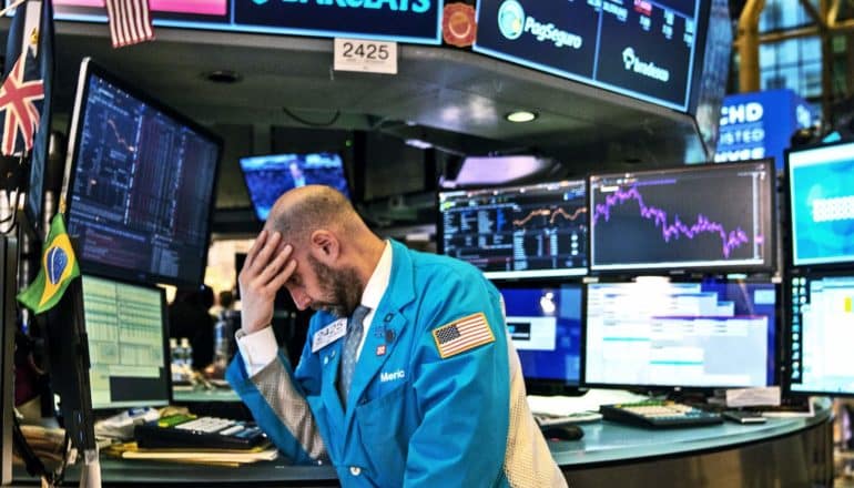 A stock market trader puts his head on his hand as screens show stock updates behind him