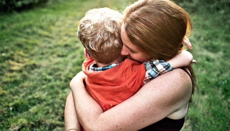 A mother hugs her sun as the sun sets with a grassy lawn in the background behind them