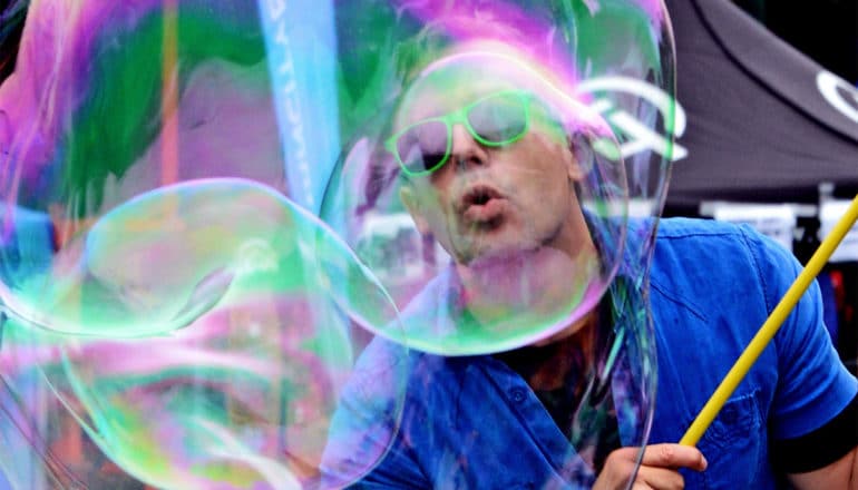 A man wearing a blue shirt and green sunglasses creating a giant soap bubble blows into the bubble