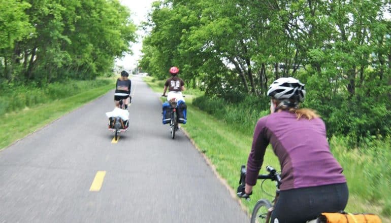 3 bikers use a paved trail through some green trees on a greenway