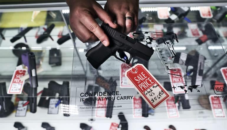 A gun shop owner holds a black pistol with a red sale tag on it over a glass case containing many pistols