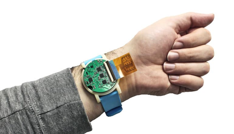 A person wears the watch-sized health monitor on their wrist. The device looks like a round, green microchip with a blue band, and an orange film touching the skin under the band.