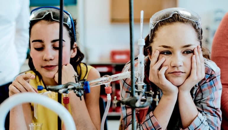 Two high school students in chemistry class look bored, one playing with a dropper and the other with her head resting on her hands