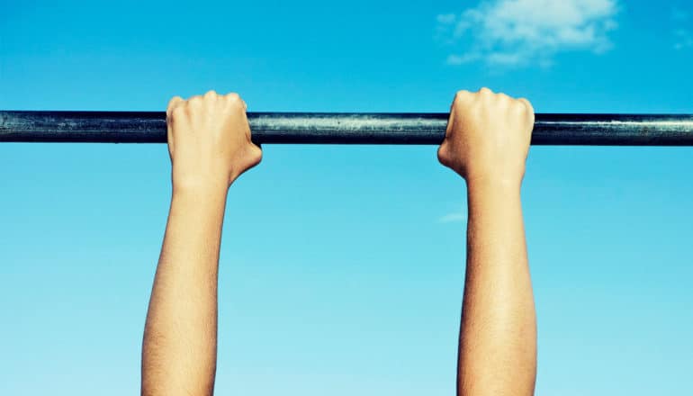 A pair of teen arms hang from a black metal pull-up bar with blue sky in the background