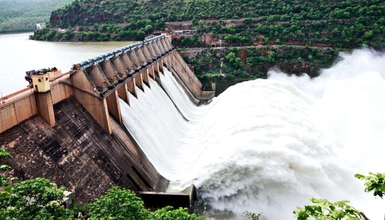 White water rushes through a hydropower dam set between green, forested mountains