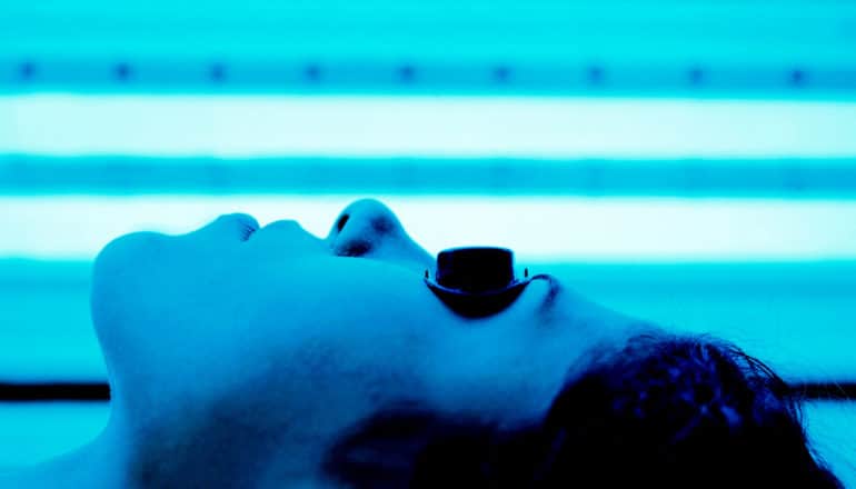 A woman lies in an indoor tanning bed, with her bathed in blue light, while wearing black goggles over her eyes