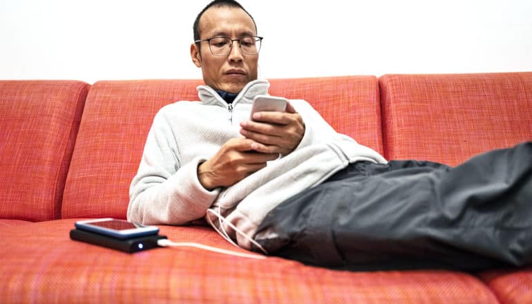 A man uses his phone on an red/orange couch while it's connected to an external battery