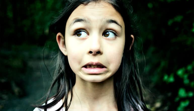 A young girl looks to her right in fear as she stands in the woods