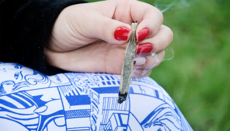 A woman with a black sleeve and blue and white pants holds a joint between fingers with red nails