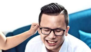 A man with glasses grimaces in pain as someone off camera pinches his ear