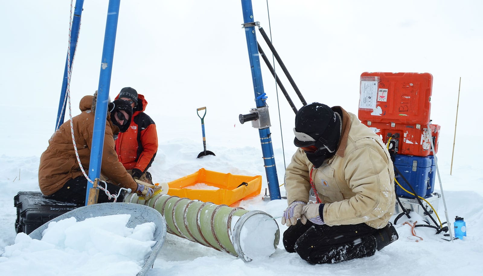 3 people in snow gear on ground with tube full of ice/snow