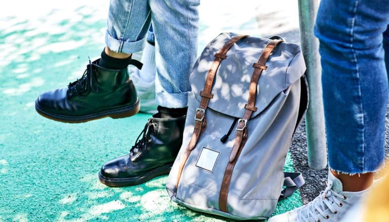 A young person in jeans and black boots dangles their legs from a railing they're sitting on, which their backpack is resting against