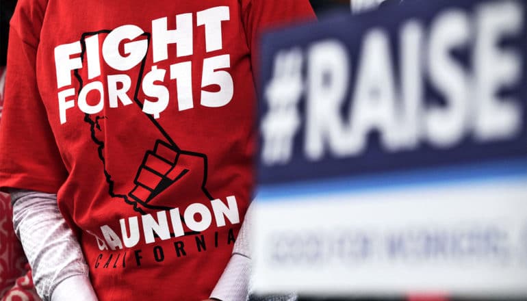 A person wears a red t-shirt that says "Fight for $15" with a picture of a raised fist over the state of California. Another person holds a sign that reads, "#RAISE."