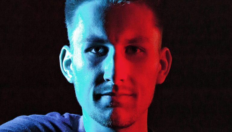 A young man looks at the camera with half his face blue and half red, with a black background