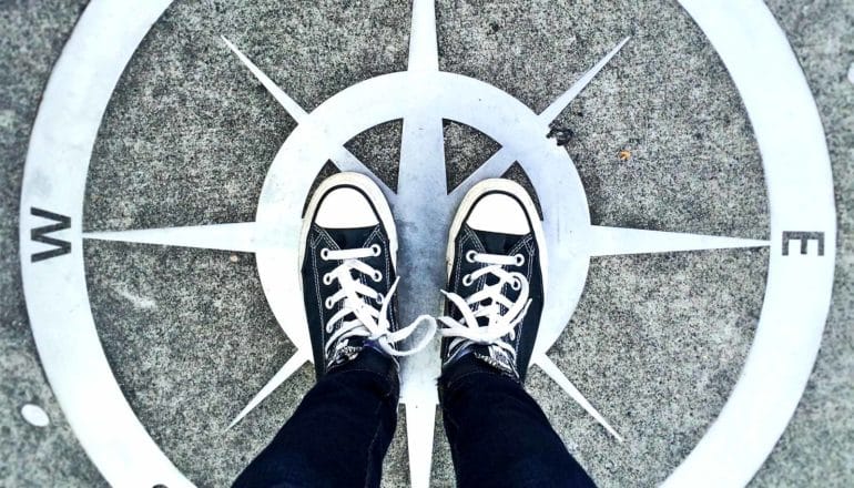 A person in dark jeans and black and white shoes stands in the middle of a compass painted on concrete