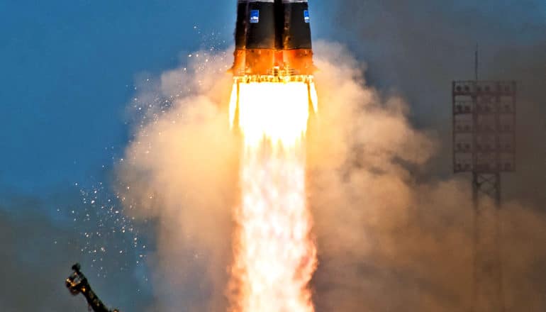 A rocket lifts off, with only its back end and the flame coming from the rocket visible. Blue sky in the background with lots of smoke from the launch