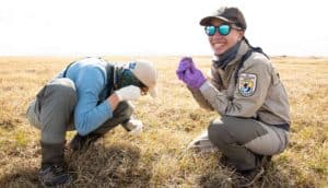 two people in field gear crouch in field. One listens to an egg, the other holds an egg and smiles