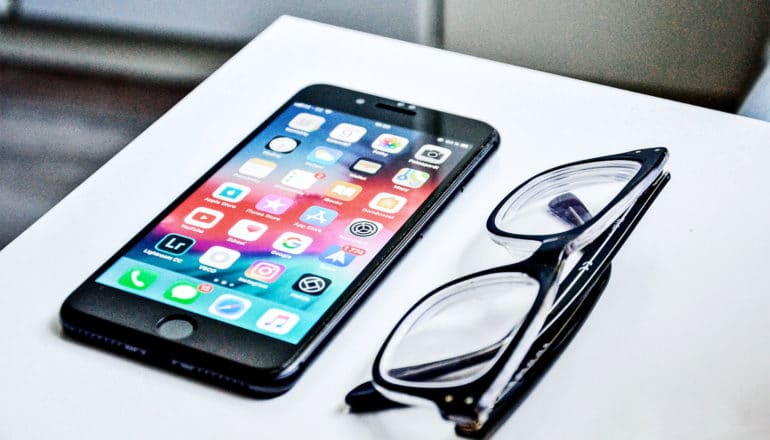 A phone sits on a white table next to a pair of black glasses