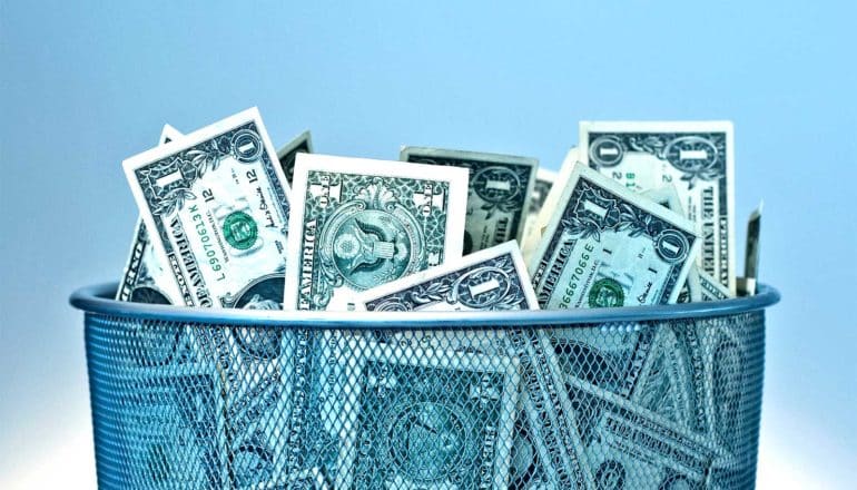 A bunch of 1 dollar bills sit in a trash can, with blue in the background