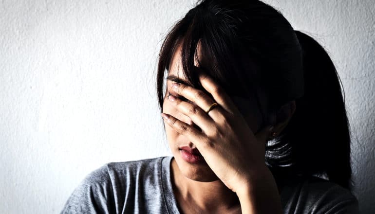 A young woman puts her hand over her face against a white wall