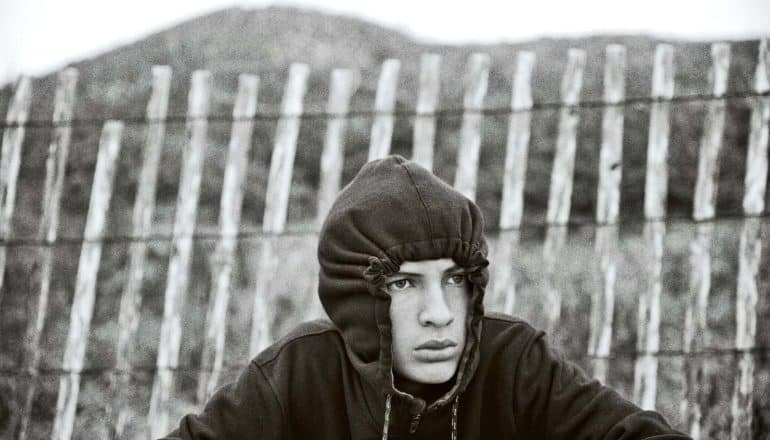 A teen boy sits near a wooden fence, with his black hoodie pulled tight around his face