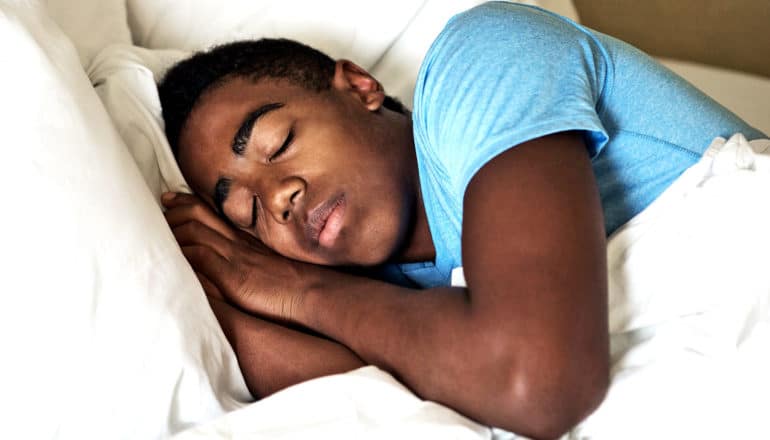 A teen boy in a blue t-shirt sleeps in bed