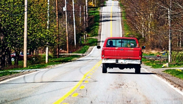 A red truck drives down a rural road, with a few houses on either side