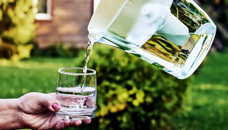 A person's hand holds out a water glass as another person off camera pours water from a pitcher with a filter