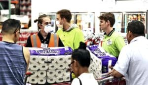 Costco workers, some of whom are wearing masks, help customers with large packages of toilet paper