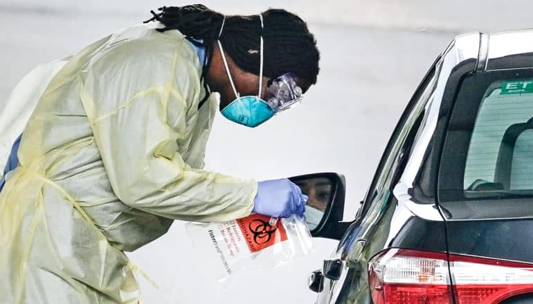 A health worker leans over a car window in full protective gear while testing for COVID-19