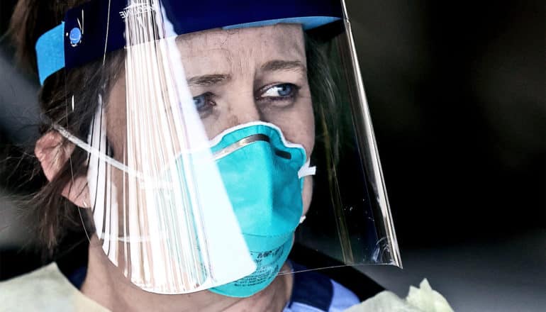 Canova wears a blue face mask and clear protective face shield looks concerned