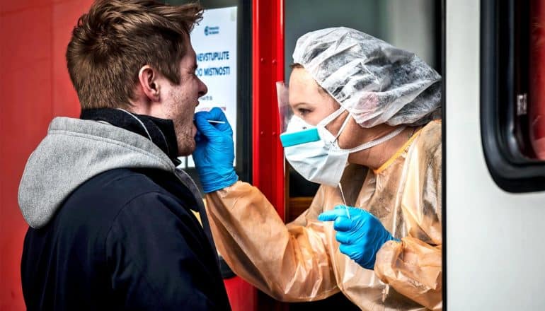 A person in medical protective gear including gloves, a mask, and a hair net, swabs a man for COVID-19