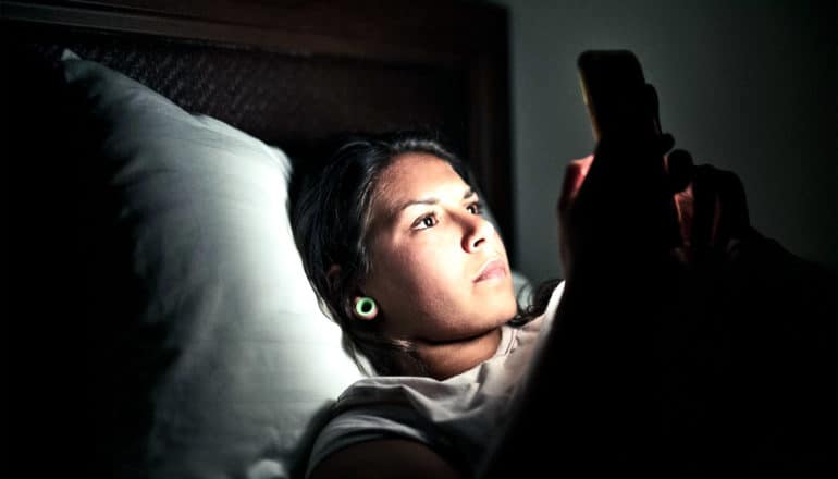 A young woman reads her phone while in bed with the lights off
