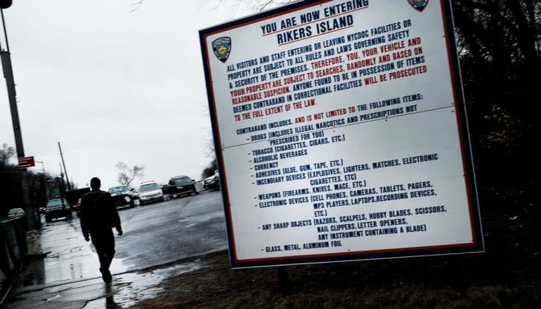 sign says "you're now entering Rikers Island" and lists contraband items. Person in silhouette walks by