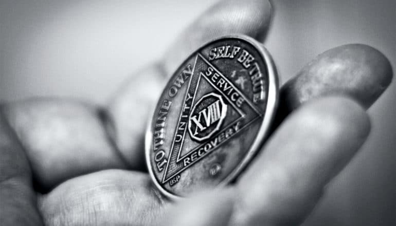A person holds their sobriety chip from Alcoholics Anonymous which shows 18 years sober. It has text that reads, "To Thine Own Self Be True" and "Unity. Service. Sobriety."