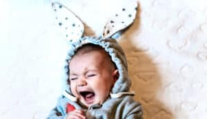A baby in a onesie with bunny ears on its hood screams while in a crib with a white pad