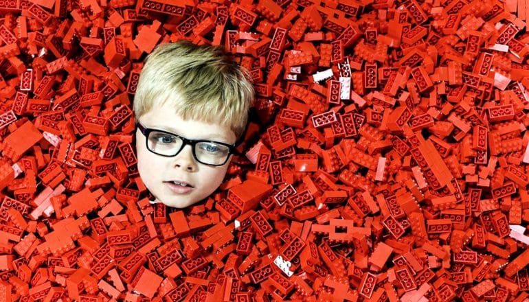 A young boy in glasses pokes his head through a sea of red Lego bricks