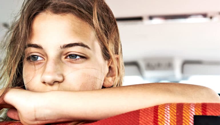 A young woman rests her face on her arm while riding in a car looking out the back window