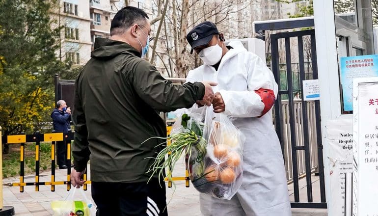 A guard in a face mask and protective suit checks a man's groceries at a gate