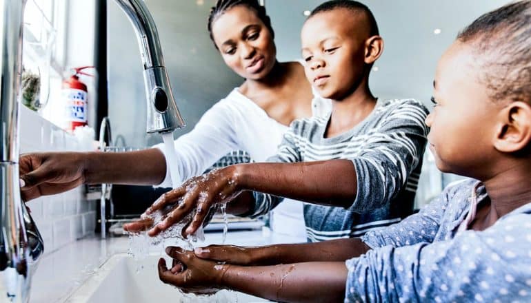 A mother helps her two children wash their hands in the kitchen sink