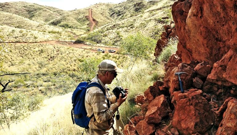 Johnson, wearing a cap, collared shirt, and backpack, looks down at red rocks with a pick axe sitting on them. A road with a few cars on it stretches into the background