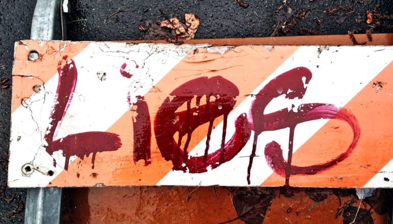 A road-block sign with orange and white stripes has the word "Lies" spray painted on it in red